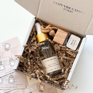 May include: A gift box from Cozy Vibe Candle Studio containing a car air freshener refill, a wooden block, and a small bottle with a dropper. The box includes instructions and is filled with brown paper. The bottle is labeled "Milk & Cereal".