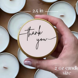 May include: A round gold metal candle tin with a light pink label featuring the black cursive text thank you. The tin has a diameter of 6.35 cm and a capacity of 57 grams. The candle contains white wax and a centered wooden wick.