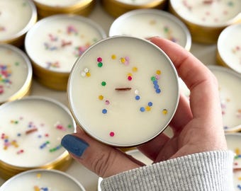 May include: Small, round candles in gold tins. The creamy white wax is decorated with colourful sprinkles. A hand holds one candle, showing the details. The candles are ideal for home fragrance.