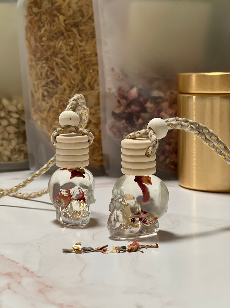 May include: Two clear glass skull-shaped bottles containing dried flowers and gold flakes. The bottles are suspended from a beige and gold cord with wooden beads. The bottles are resting on a white marble surface.