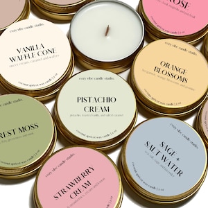 May include: Assortment of 10 small, round, gold-rimmed candle jars with lids. Each jar has a different scent and label. The scents include: Vanilla Waffle Cone, Pistachio Cream, Strawberry Cream, Orange Blossoms, Sage + Salt Water, and Rest Moss. Each jar is labeled with the scent and a short description of the scent.