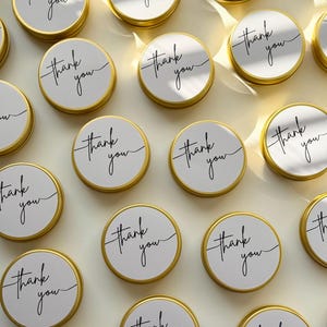 May include: A collection of round, gold-coloured metal tins arranged in a grid. Each tin features a white circular label on the lid with the words thank you printed in a black, cursive script font. The lids have a smooth, metallic finish.