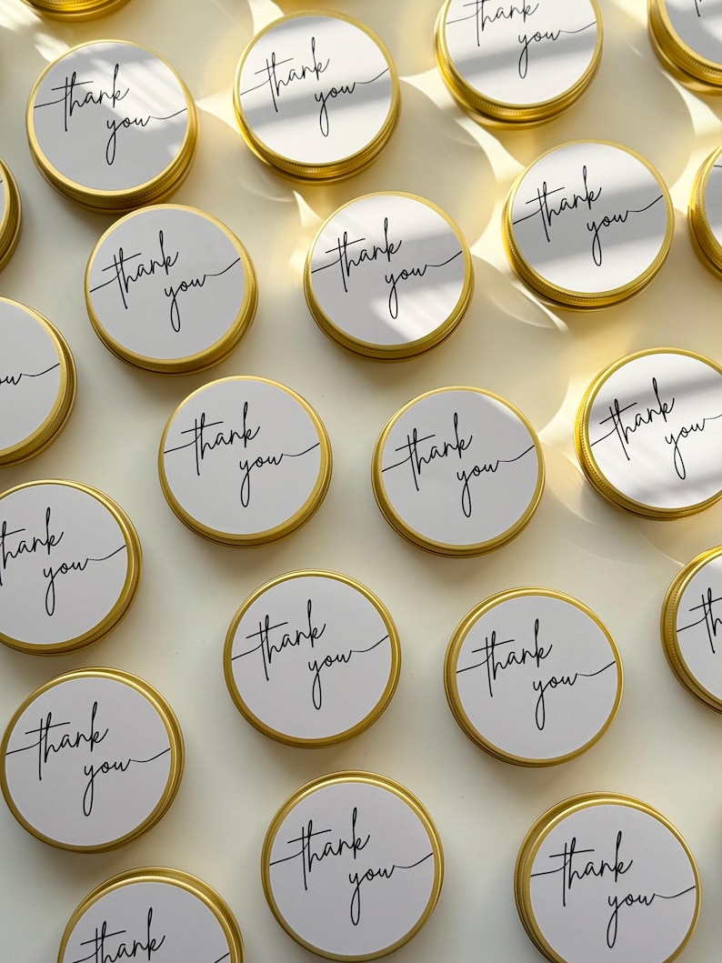May include: A collection of round, gold-coloured metal tins arranged in a grid. Each tin features a white circular label on the lid with the words thank you printed in a black, cursive script font. The lids have a smooth, metallic finish.