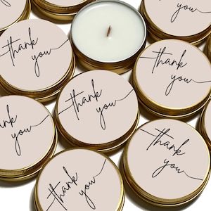 May include: A collection of round gold metal tins with lids featuring a light beige label printed with the words thank you in a black cursive script. One tin is open, revealing a white wax candle with a single centred wooden wick.