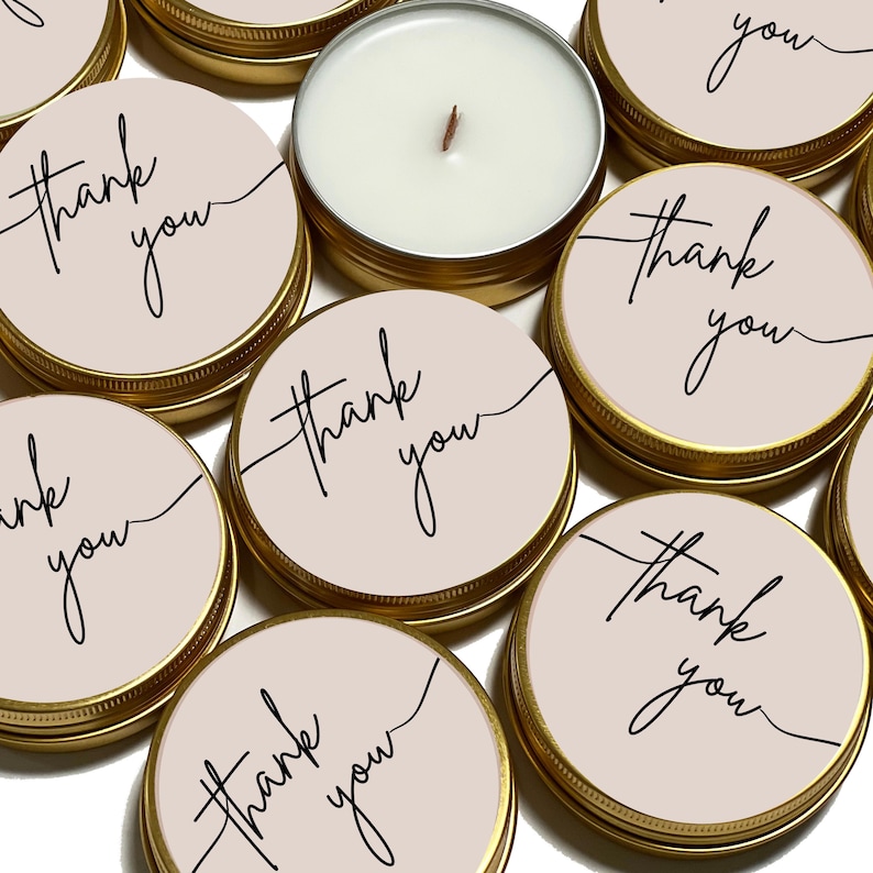 May include: A collection of round gold metal tins with lids featuring a light beige label printed with the words thank you in a black cursive script. One tin is open, revealing a white wax candle with a single centred wooden wick.
