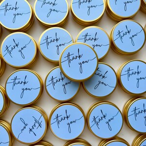 May include: A collection of round gold metal tins with light blue lids. Each lid features the text thank you written in a black cursive script. The tins are arranged in a cluster, showcasing the repeating design across the circular surfaces.