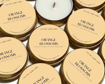 May include: A collection of small, gold-rimmed candle jars with a light yellow label. The label reads "ORANGE BLOSSOMS" with the text "bergamot, orange blossoms and jasmine" underneath. Each jar contains a white candle and has a label that reads "coconut apricot wax candle 1.2 oz" and "cozy vibe candle studio."
