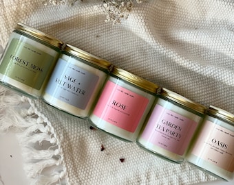 May include: Five glass candle jars with gold lids and labels. The candles are labeled "Forest Moss", "Sage + Saltwater", "Rose", "Garden Tea Party", and "Oasis". Each jar contains 24 ml of candle wax.