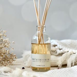 May include: Clear glass reed diffuser bottle filled with a light amber liquid and wooden reeds. The label reads "Pistachio Cream" with details of pistachio, toasted vanilla, and salted caramel. The bottle is 100 ml.