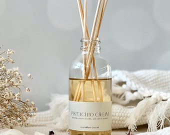May include: A clear glass reed diffuser bottle containing a pale amber liquid and wooden reeds. The label reads "Pistachio Cream" with details of pistachio, toasted vanilla, and salted caramel. The bottle is 100 ml.
