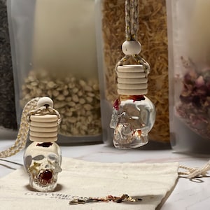 May include: Two clear glass skull-shaped bottles with a red floral design inside. The bottles are hanging from a beige cord with wooden beads. The bottles are sitting on a white fabric surface with the text "Cozy Vibe Candles" printed on it.