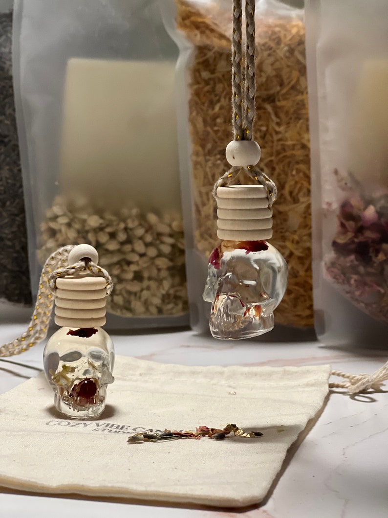May include: Two clear glass skull-shaped bottles with a red floral design inside. The bottles are hanging from a beige cord with wooden beads. The bottles are sitting on a white fabric surface with the text "Cozy Vibe Candles" printed on it.