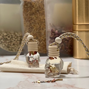 May include: Two clear glass skull-shaped bottles containing dried flowers, suspended from a gold cord with white wooden beads. The bottles are positioned on a white marble surface.