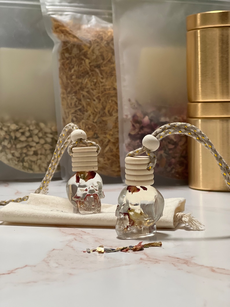 May include: Two clear glass skull-shaped bottles containing dried flowers, suspended from a gold cord with white wooden beads. The bottles are positioned on a white marble surface.