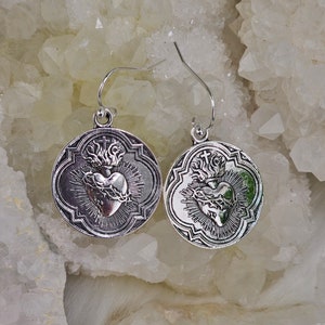 May include: Silver earrings with a detailed design of a heart with a crown of thorns and flames. The heart is surrounded by a decorative frame.