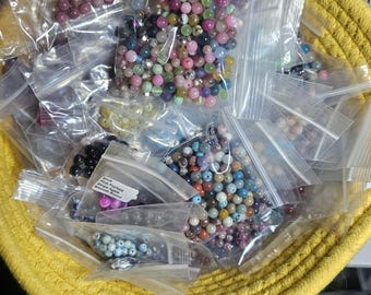 Puede incluir: Una colección de pequeñas bolsas de plástico transparente resellables llenas de cuentas redondas de piedras preciosas en varios colores, incluyendo rosa, azul, verde y negro, contenidas en una cesta redonda de mimbre amarillo con dos asas.