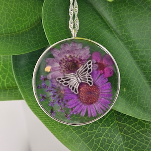 May include: A silver chain necklace with a round pendant featuring a pressed purple flower and a silver butterfly charm.