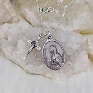 May include: A silver chain necklace with two pendants. One pendant is a silver cross with a figure of Jesus Christ. The other pendant is a silver oval with a depiction of Saint Monica. The necklace is displayed on a white background.