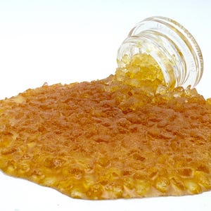 Op de afbeelding: Een heldere glazen pot die is omgekieperd en een gouden, kristallijne substantie op een wit oppervlak morst. De substantie vormt een getextureerde, amberkleurige plas. De deksel van de pot is zichtbaar.