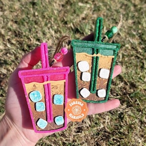 May include: Two iced coffee ornaments, one pink and one green, with white marshmallows and blue ice cubes. The ornaments are made of wood and have a glitter finish. The ornaments are hanging from a string with beads. The ornaments are on a hand with a "Forever Marigold" sticker on the hand.