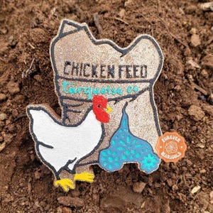 May include: A decorative pin shaped like a sack labeled CHICKEN FEED in black text with Turquoise Co written below in teal. A white chicken with a red comb and yellow feet stands in front of the sack next to a blue patterned shape.