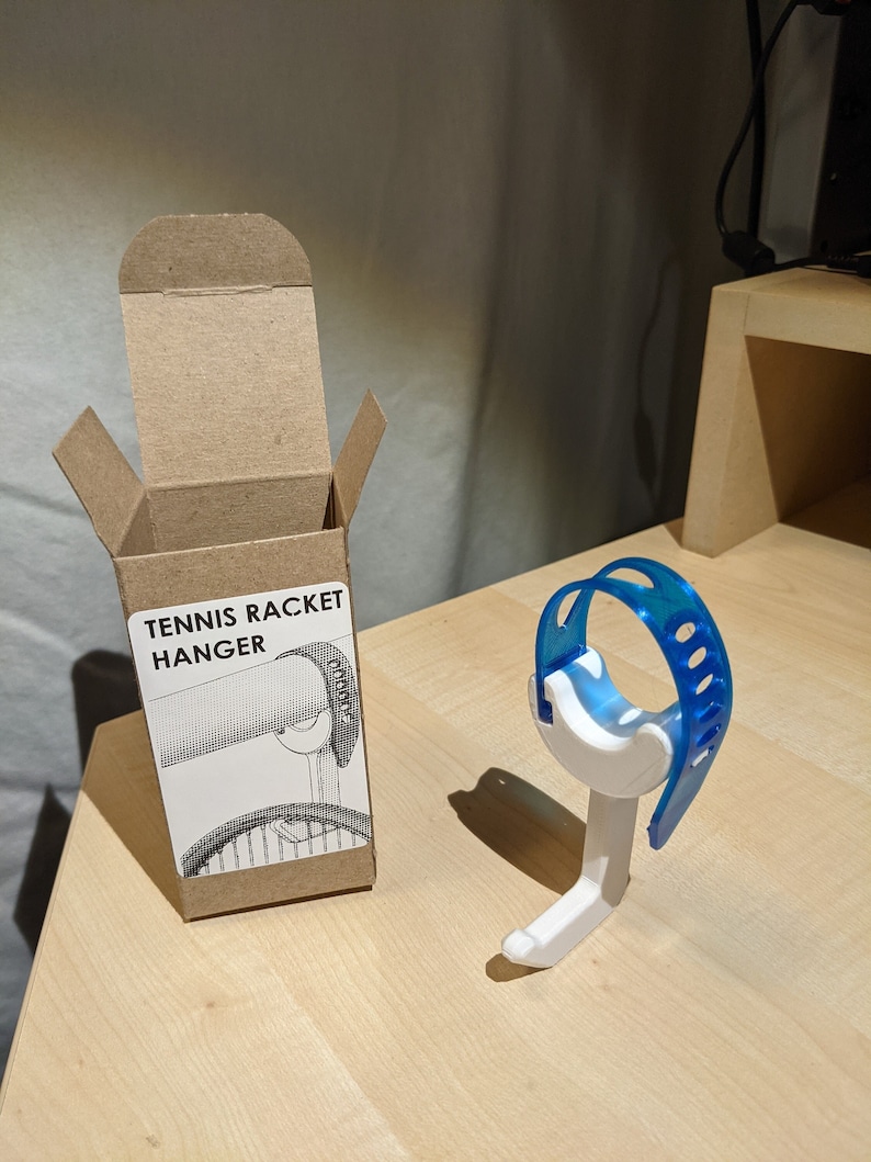 Puede incluir: Un colgador de raqueta de tenis de pl&aacute;stico blanco con una correa de pl&aacute;stico azul. El colgador est&aacute; empaquetado en una caja de cart&oacute;n marr&oacute;n con el texto "Tennis Racket Hanger" impreso en la parte delantera.
