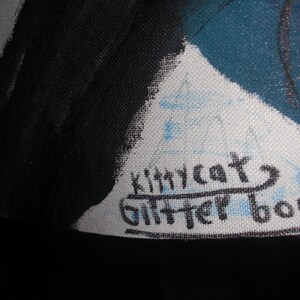 May include: Close-up of a canvas with a painting featuring black and teal colors. The artist's signature, "kittycat glitter boom," is visible in black ink on a white section of the canvas. The canvas is stretched over a wooden frame.