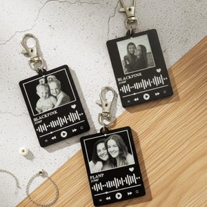 May include: Three black keyrings with silver clasps, each displaying a grayscale photo and a music waveform design. The keyrings have the text "BLACKPINK JUMP" and "PLANP JUMP". They are arranged on a wooden surface.