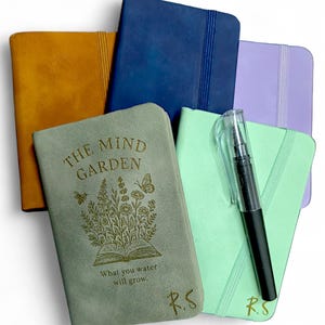 May include: A collection of small notebooks in colours such as mustard, navy, and mint green. One notebook has the text "THE MIND GARDEN" with a floral design and the quote "What you water will grow." A black pen is placed on one of the notebooks.
