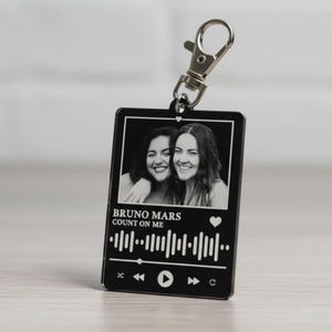 Puede incluir: Llavero rectangular negro con un cierre plateado. El llavero presenta una foto en blanco y negro de dos personas sonriendo. Debajo de la foto est&aacute; el texto "BRUNO MARS COUNT ON ME" y una forma de onda musical.