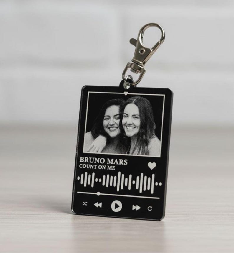 Puede incluir: Llavero rectangular negro con un cierre plateado. El llavero presenta una foto en blanco y negro de dos personas sonriendo. Debajo de la foto est&aacute; el texto "BRUNO MARS COUNT ON ME" y una forma de onda musical.