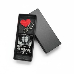 Puede incluir: Un llavero acr&iacute;lico negro con una foto de dos personas, una forma de onda musical y el texto "BRUNO MARS COUNT ON ME". El llavero est&aacute; en una caja negra abierta, con un coraz&oacute;n rojo y un cierre plateado.