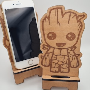 May include: Wooden phone stand featuring a cartoon image of Groot from Guardians of the Galaxy. The stand is brown and has a slot for a phone.