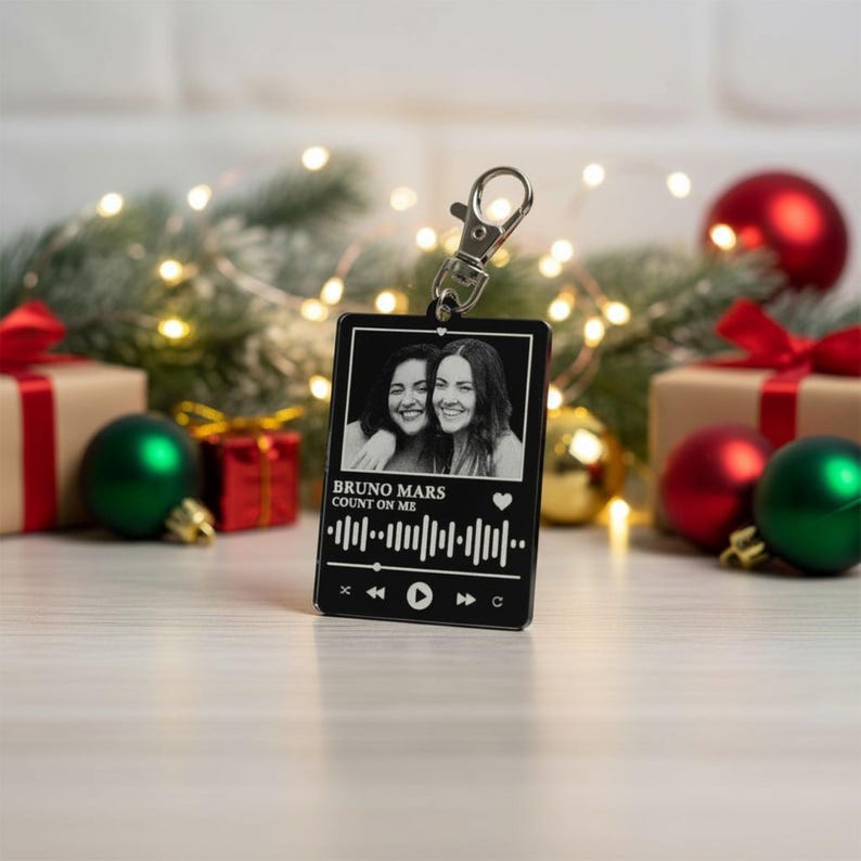 Puede incluir: Un llavero negro con un cierre plateado, con una foto en blanco y negro de dos personas sonriendo. El llavero tiene el texto "BRUNO MARS COUNT ON ME" y un gr&aacute;fico de forma de onda musical. Decoraciones festivas navide&ntilde;as en el fondo.