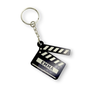 May include: A black and white film clapperboard keyring with the name "EMMA" printed on it. The keyring has a silver ring and chain. The clapperboard design is a classic movie symbol.