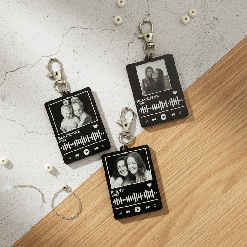 Puede incluir: Tres llaveros negros con cierres plateados, cada uno con una foto en escala de grises y un dise&ntilde;o de forma de onda musical. Los llaveros tienen el texto "BLACKPINK JUMP" y "PLANP JUMP". Est&aacute;n sobre una superficie de madera.