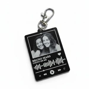 Puede incluir: Llavero rectangular negro con un cierre plateado. El llavero presenta una foto en blanco y negro de dos personas sonriendo. Debajo de la foto est&aacute; el texto "BRUNO MARS COUNT ON ME" y un gr&aacute;fico de reproductor de m&uacute;sica.