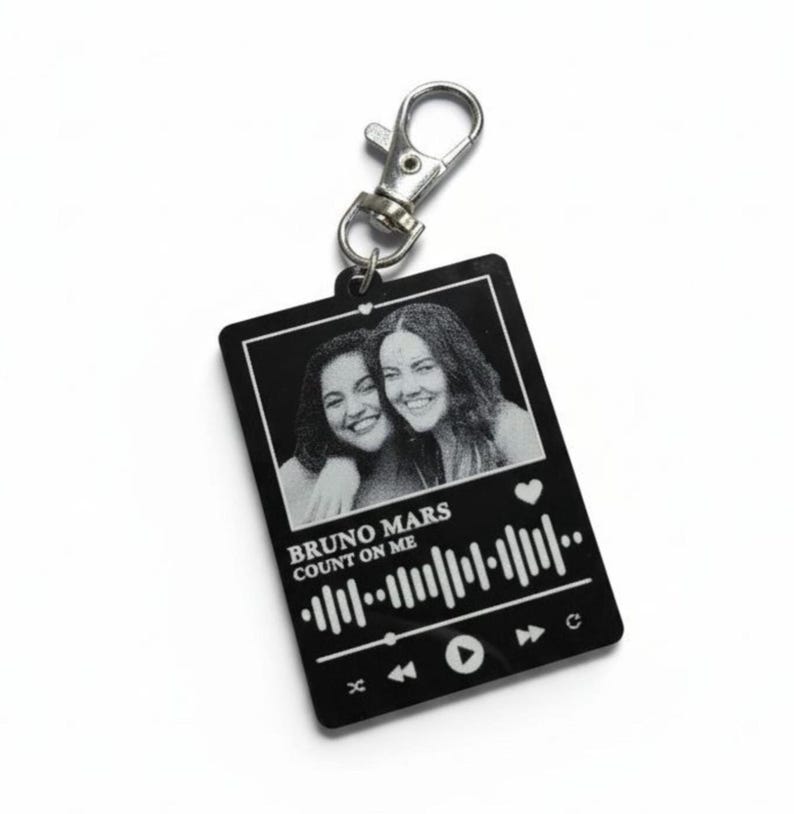 Puede incluir: Llavero rectangular negro con un cierre plateado. El llavero presenta una foto en blanco y negro de dos personas sonriendo. Debajo de la foto est&aacute; el texto "BRUNO MARS COUNT ON ME" y un gr&aacute;fico de reproductor de m&uacute;sica.