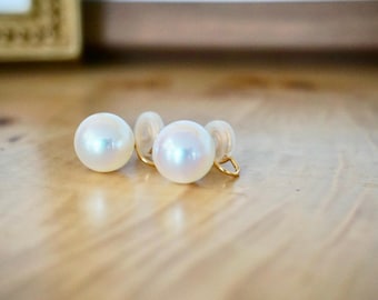 May include: Two white pearl earrings with gold-colored clasps. The earrings are round and have a lustrous, iridescent sheen. They are resting on a light-colored wooden surface, with a blurred background.