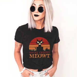 May include: Black t-shirt featuring a retro design with a black cat silhouette against a sunset backdrop. The text "YOU'RE CREEPIN MEOWT" is printed below the cat. A fun and spooky design for Halloween.