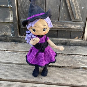 Pode incluir: Uma boneca de bruxa de croch&ecirc; com cabelo roxo, um chap&eacute;u preto e um vestido roxo. A boneca est&aacute; em p&eacute; sobre uma superf&iacute;cie de madeira.