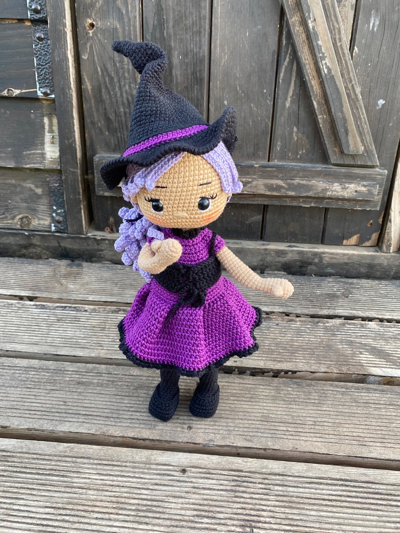 Pode incluir: Uma boneca de bruxa de croch&ecirc; com cabelo roxo, um chap&eacute;u preto e um vestido roxo. A boneca est&aacute; em p&eacute; sobre uma superf&iacute;cie de madeira.