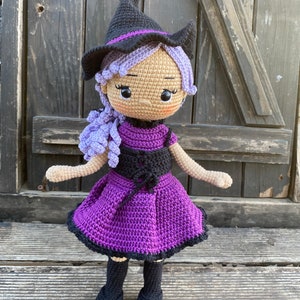 Pode incluir: Uma boneca de bruxa de croch&ecirc; com cabelo roxo, um chap&eacute;u preto e um vestido roxo. A boneca est&aacute; em p&eacute; sobre uma superf&iacute;cie de madeira.