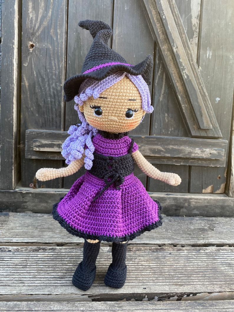 Pode incluir: Uma boneca de bruxa de croch&ecirc; com cabelo roxo, um chap&eacute;u preto e um vestido roxo. A boneca est&aacute; em p&eacute; sobre uma superf&iacute;cie de madeira.