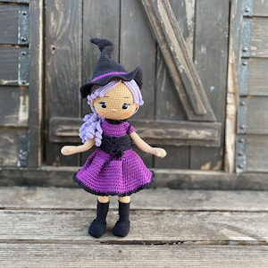 Pode incluir: Uma boneca de bruxa de croch&ecirc; com cabelo roxo, um chap&eacute;u preto e um vestido roxo. A boneca est&aacute; em p&eacute; sobre uma superf&iacute;cie de madeira.