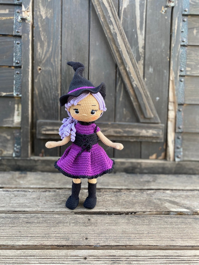 Pode incluir: Uma boneca de bruxa de croch&ecirc; com cabelo roxo, um chap&eacute;u preto e um vestido roxo. A boneca est&aacute; em p&eacute; sobre uma superf&iacute;cie de madeira.