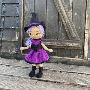 Pode incluir: Uma boneca de bruxa de croch&ecirc; com cabelo roxo, um chap&eacute;u preto e um vestido roxo. A boneca est&aacute; em p&eacute; sobre uma superf&iacute;cie de madeira.