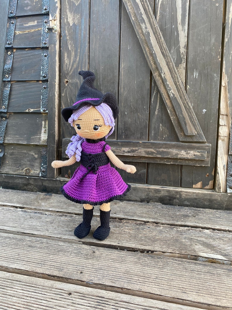 Pode incluir: Uma boneca de bruxa de croch&ecirc; com cabelo roxo, um chap&eacute;u preto e um vestido roxo. A boneca est&aacute; em p&eacute; sobre uma superf&iacute;cie de madeira.