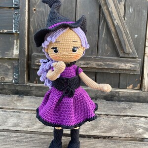 Pode incluir: Uma boneca de bruxa de croch&ecirc; com cabelo roxo, um chap&eacute;u preto e um vestido roxo. A boneca est&aacute; em p&eacute; sobre uma superf&iacute;cie de madeira.