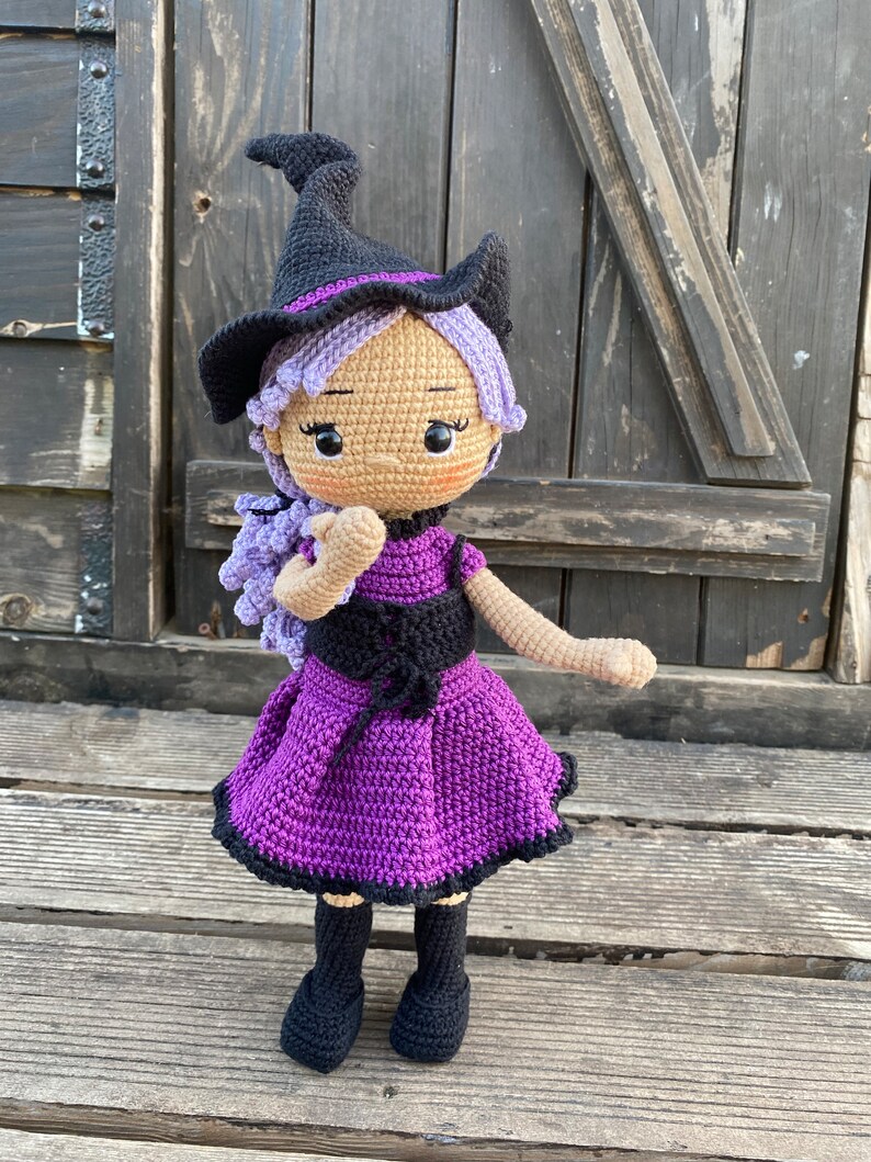Pode incluir: Uma boneca de bruxa de croch&ecirc; com cabelo roxo, um chap&eacute;u preto e um vestido roxo. A boneca est&aacute; em p&eacute; sobre uma superf&iacute;cie de madeira.